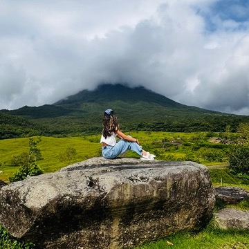 Me sitting on a big rock in Costa Rica, look at a volcano covered in clouds and surrounded by green hills.